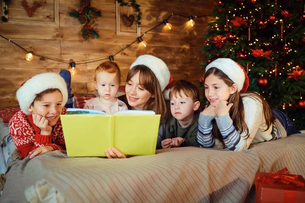 Mor läser en bok med barn liggande på sängen i rummet med träd på julen. — Stockfoto