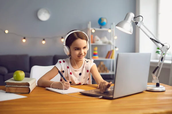 Домашня онлайн-школа. Працьовита маленька дівчинка в гарнітурі дивиться онлайн урок на ноутбуці — стокове фото