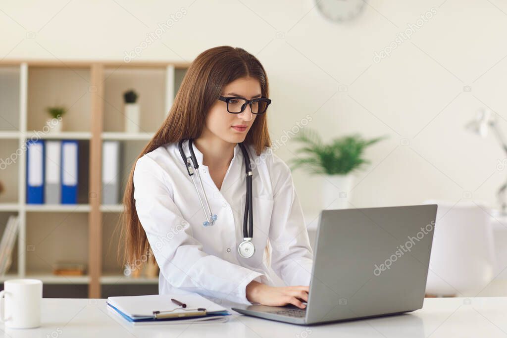 Young female doctor typing on laptop keyword surfing Internet in office of modern clinic