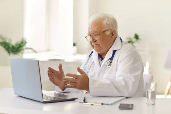 Ernstige senior arts zit aan het bureau in het ziekenhuis kantoor en met behulp van laptop om zijn patiënten te bellen video — Stockfoto