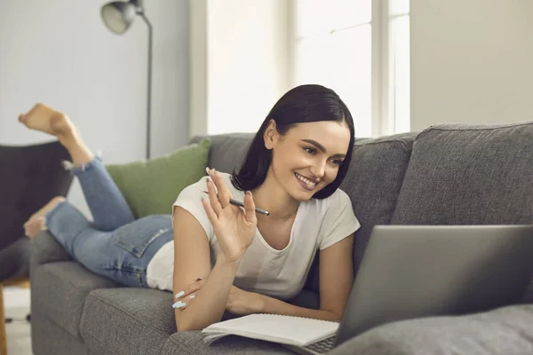 Smiling woman lying on sofa with laptop and greeting somebody with hand during videocall — Stock Photo, Image