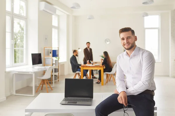 Joven hombre de negocios con portátil con pantalla en blanco sentado en el escritorio en el espacio de coworking mirando a la cámara — Foto de Stock