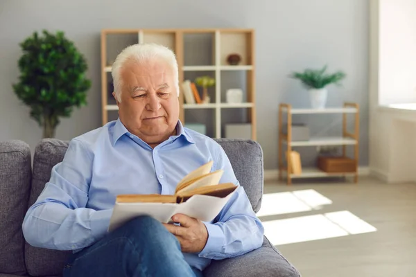 Щасливий пенсіонер проводить дозвілля вдома і читає стару книгу, сидячи на дивані у вітальні — стокове фото