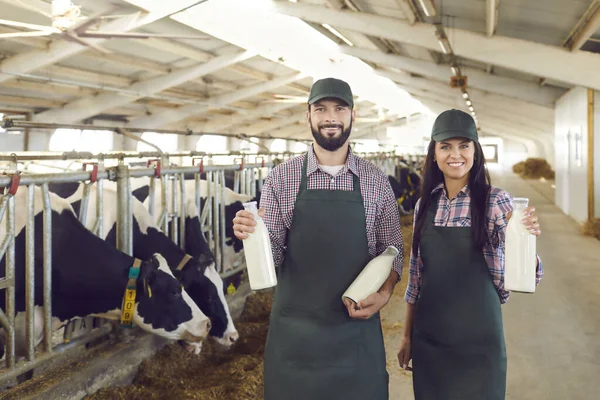 Felices propietarios de granjas lecheras de pie en un establo, sosteniendo botellas de leche, sonriendo y mirando a la cámara —  Fotos de Stock