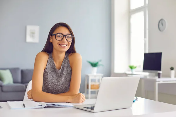 Счастливая молодая женщина в крутых очках сидит за столом с открытым ноутбуком и ноутбуком, глядя в камеру — стоковое фото