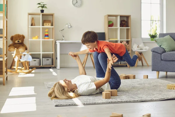 Happy young mother lying on carpet, holding her son on legs in raised hands and having fun at home — Stock Photo, Image