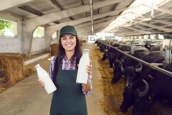 Mujer joven feliz de pie en el establo, sosteniendo botellas de leche fresca y mirando a la cámara —  Fotos de Stock