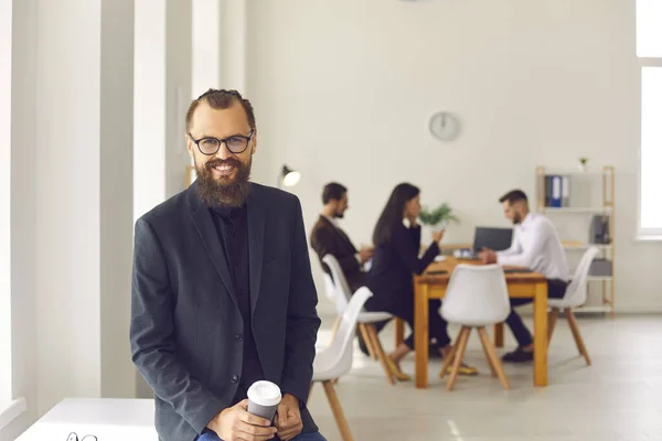 Glad ung affärsman med kaffe att gå i moderna coworking utrymme med upptagen människor i bakgrunden — Stockfoto