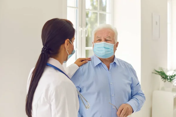 Starszy pacjent i młoda lekarka, nosząca maski medyczne, komunikująca się w gabinecie szpitalnym — Zdjęcie stockowe