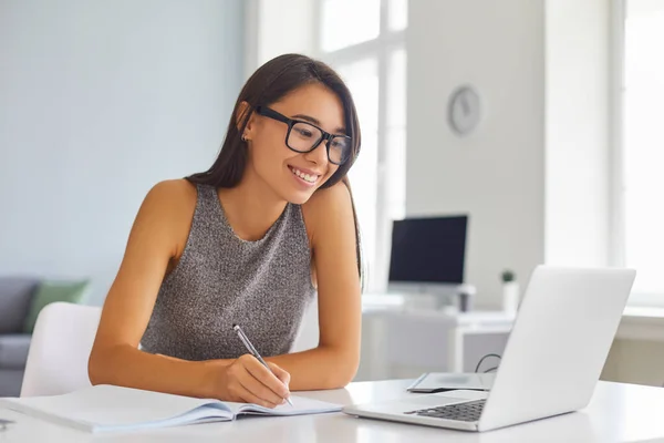 Улыбающаяся офисная работница сидит за столом, смотрит на экран ноутбука и делает заметки во время онлайн-конференции — стоковое фото