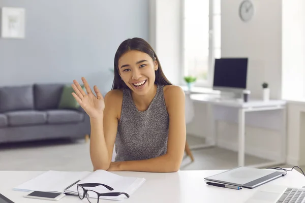 Smiling asian woman looking at camera and greeting partner with hand during online videocall in office — Stock Photo, Image