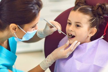 Kadın diş hekimi, diş kliniğinde gülümseyen kız dişlerini aynayla inceliyor.