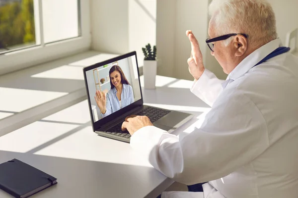 Старший лікар у формі та окулярах сидить у ноутбуці та вітає молоду усміхнену жінку-пацієнтку онлайн — стокове фото