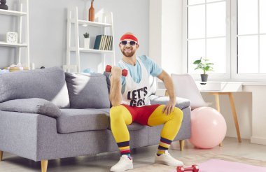 Komik adam evdeki kanepede oturup spor yapmak için dambıl egzersizi yapıyor. Bu ev çalışması oturma odasına spor salonu deneyimini getiriyor. Spor salonuna mizah katıyor..