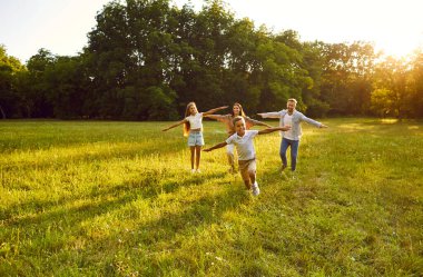 Mutlu aile koşuları ve yaz parkında eğlenmeler. Doğanın güneşli günlerinin tadını çıkarıyorlar. Anne, baba ve çocukları gün batımında uçmak ve dışarıda uçmak gibi kollarını kaldırıyorlar..