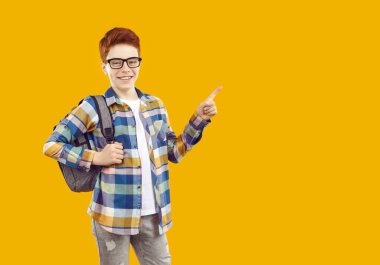 Sırtında sırt çantasıyla ekose gömlek ve kot pantolon giymiş gözlüklü mutlu, kızıl saçlı okul çocuğu izole edilmiş sarı arka plandaki boş alanı işaret ediyor. Okul eğitimi kavramı.