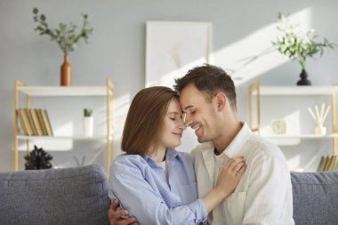 Oturma odasındaki kanepeye yakın oturan mutlu çift kucaklaşıyorlar ve birbirlerine içtenlikle gülümsüyorlar. Sevinçli bir çift birbirlerine sarılırlar, yakınlık anlarını paylaşırlar ve aralarında barışçıl bir bağ kurarlar..