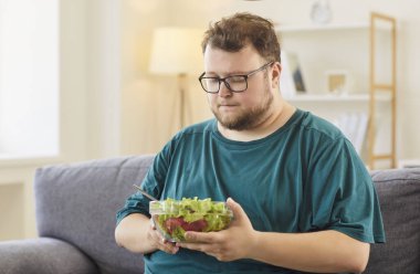Şişman şişman adam salata, diyet ve sağlığı düşünüyor. Evdeki bir kanepede, beslenme, porsiyon, seçim ve hedefleri tartıyor, yaşam tarzını değiştiriyor. Sağlıklı beslenme ve kilo kaybı.