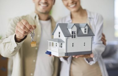 Satın alındıktan sonra ev anahtarlarını tutan mutlu çift. Emlak başarısı ve ipotek onayını simgeleyen bir model evle gülümsüyorlar. Parlak kutular istiflendi. Yeni ev sahiplerini kabul et.