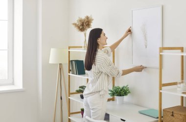 Neşeli genç bir kadın duvarda sanat eseri düzenliyor, güneşli, çağdaş dairesinde duvara resim asıyor, gülümsüyor ve düzenliyor, yaşam alanını dekore ediyor. Konut geliştirme kavramı 