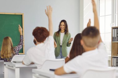 Lise öğrencileri sınıf sıralarında mutlu bir bayan öğretmenle oturuyor ve bir ders sırasında soru sormak ya da cevap vermek için ellerini kaldırıyorlar. Okula dönmek, eğitim kavramı.