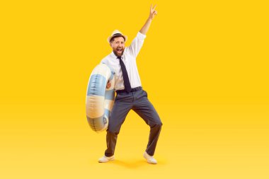 Neşeli erkek ofis çalışanı uzun zamandır beklenen yaz tatiline sonunda gittiği için mutlu. Resmi kıyafetli eğlenceli iş adamı yüzme çemberi tutuyor ve turuncu arka planda V işareti gösteriyor..