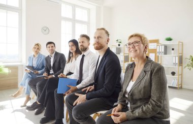 İş merkezinin ofisindeki iş eğitimi oturumuna çeşitli profesyonel gruplar katılır. Resmi giyinmiş erkek ve kadınlar sıraya geçip hoparlör ya da sunumla ilgilenirler.
