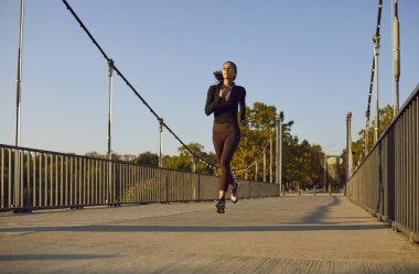 Şehir parkında köprüden koşarak geçen zayıf, genç bir kadın. Esmer kız spor giyinmiş, dışarıda koşuyor. Kilo kaybı, sağlıklı yaşam tarzı, spor antrenmanı ve doğa konseptinde uygunluk..