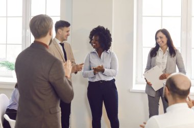 Ofis çeşitliliğindeki farklı etnik gruplardan iş arkadaşları çalışma alanında grup halinde duruyor, kurumsal başarılarını, takım çalışmalarını gösteriyorlar, yeni çalışanları sıcak bir şekilde karşılıyorlar, mülakatta başvuranları alkışlıyorlar.