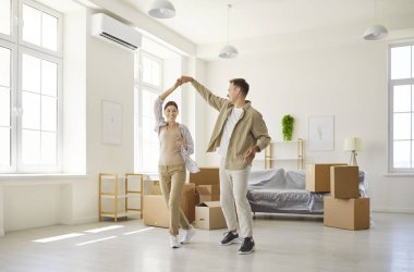 Bugün kutuları taşırken dans eden gülümseyen çift. Geniş pencereli parlak bir apartman dairesi bavulların açıldığını, eğlencenin ve takım çalışmasının paylaşıldığını gösteriyor. Tema yer değiştirme, aşk ve yeni başlangıçlar.