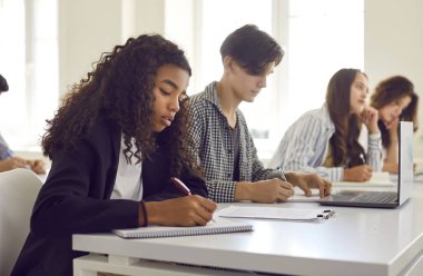 Sınıftaki bir kolej dersi sırasında sırayla masada oturan ve yazı sınavı yazan bir grup farklı lise öğrencisinin yan görüntüsü. Eğitim ve bilgi kavramı.