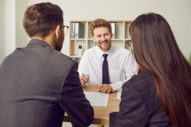 Ev almak isteyen müşterilerle arkadaş canlısı bir emlakçı toplantısı. Mutlu, gülümseyen bir adam aile ile birlikte çalışma masasında oturuyor ve onlara kağıt sözleşmesi ve imzalamaları için bir kalem veriyor.