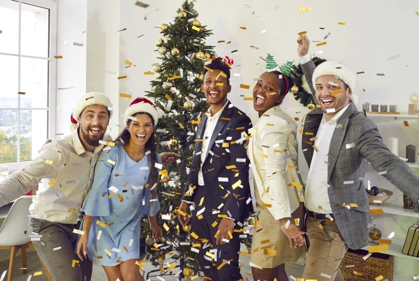 Neşeli farklı arkadaşlar Noel partisinde eğleniyorlar. Noel Baba şapkalı mutlu ve heyecanlı genç insanların grup portresi süslenmiş odada altın konfetiyle dikiliyor.