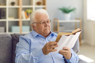 50-60 yaşlarında, ciddi derecede zeki beyaz saçlı bir adam oturma odasında kanepede oturuyor, boş zamanlarını sessizce geçirmek için kitap okuyor, en çok satan kitaplardan keyif alıyor, genç kalmayı ve yeni gözlükleri test ediyor.