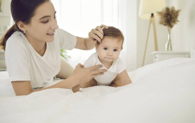Anne ve çocuk beyaz yatakta. Anne ve altı aylık bebek, güneşli yatak odasında rahatlıyor, oynuyor ve birlikte eğleniyorlar. Yatakta uzanan anne, kameraya bakan bebek saçlarını nazikçe düzeltir.