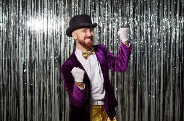 Heyecanlı adam canlı mor bir kostüm içinde, beyaz eldivenler ve silindir şapkayla zaferi kutluyor. Yumruklarını sıkmış, parıldayan gümüş renkli stüdyo arka planında duruyor.