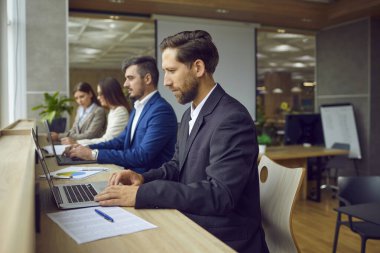 Ciddi iş adamları, dizüstü bilgisayarda oturup modern açık planlı ofiste aynı masada oturuyorlar. Şirket çalışanları iş yerlerinde bilgisayar yazarken takım elbise giyiyorlar..