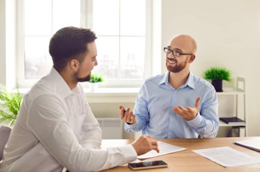İş yerindeki işadamları. İki genç, gülümseyen işveren işyeri masasında oturuyor, konuşuyor ve tartışıyorlar, yeni projeleri veya girişimleri değerlendiriyorlar, şirketi analiz ediyorlar..