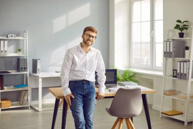 Ofiste başarılı bir iş adamının portresi. Beyaz gömlekli, kot pantolonlu ve gözlüklü yakışıklı iş adamı çalışma masasının yanında duruyor.