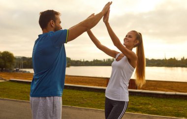 Erkek koç ya da antrenör, şehir parkında spor yaparken spor eğitimi alan mutlu stajyer bir kadına beşlik çakıyor. Zindelik yolculuğunda beraberlik teşvik ve destek.