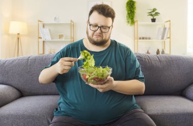 Evdeki kanepede salata yiyen mutsuz şişman şişman adam. Elinde bir kase yeşillik var, diyet konusunda isteksiz ve evde daha sağlıklı oturuyor. Sağlıklı beslenme, yemek, yaşam tarzı ve alışkanlıklar değişir..
