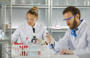 Tıbbi biyoteknoloji kimya laboratuvarında çalışma süreci. Kadın bilim adamı mikroskop altında mikroorganizmaları incelerken erkek meslektaşı da pipetle örnek alıyor. Seçici odak.