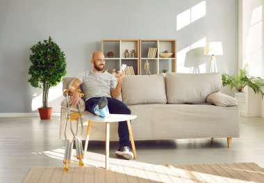 Bacağından yaralanan adam arkadaşlarıyla çevrimiçi iletişim kurmak ya da sosyal ağları taramak için cep telefonu kullanıyor. Koltukta oturan gülümseyen genç adam koltuk değneklerinin yanında oturuyor ve telefon ekranına bakıyordu..