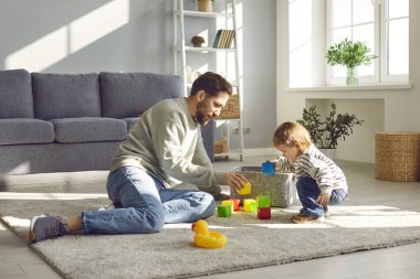 Ebeveynlik. Sevgi dolu genç bir baba ve küçük kızı evde renkli bloklarla oynuyorlar. Beyaz bir baba oturma odasında oturmuş küçük çocuğuyla birlikte hafta sonunun tadını çıkarıyor..