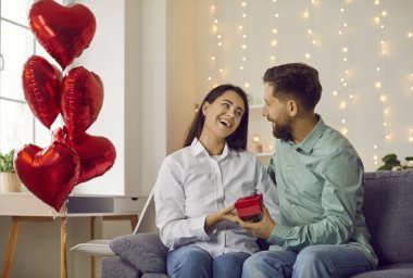 Sevgililer Günü 'nü kutlayan sevimli adam ve kadın. Mutlu çift hediye kutusunu bir arada tutarken birbirlerine şefkatle bakarken kalp şeklinde balonların arka planında koltukta oturuyorlar.