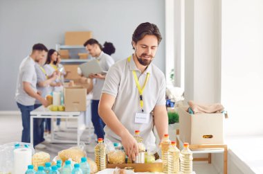 Yemek bankasında gönüllü çalışıyor. Üniformalı genç bir erkek işçi. Üzerinde kimlik etiketi var. Yağ, makarna ve diğer yiyecekleri kutulara koyuyorlar. Gıda bağışı, açlık yardımı, insani yardım, hayır işi kavramları