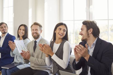 Ofis toplantısı ya da etkinlik sırasında neşeli ve gülümseyen çeşitli iş insanları alkışlıyor. Çalışma ortamında takım çalışması, başarı ve profesyonel işbirliği vurgulandı.