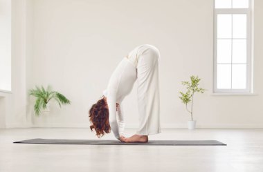 Genç, sıska kadın yoga yapıyor, öne eğilip egzersiz yapıyor. Hafif stüdyoda uttanasana pozunda beyaz spor giysili bir kadın. Spor, spor ve antrenman konsepti.