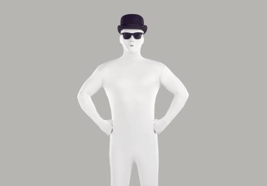 Siyah şapkalı ve gözlüklü, beyaz taytlı, tanınmayan birinin portresi. Vücut kıyafetli adam, gri arka planda izole edilmiş bir şekilde stüdyoda kameraya kendinden emin bir şekilde bakıyor..