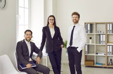 Üç kişilik bir iş adamının portresi kameraya bakıyor ve işyerinde gülümsüyor. Şirket çalışanları ya da çalışanlar ofiste güvenle ve neşeyle duruyorlar..